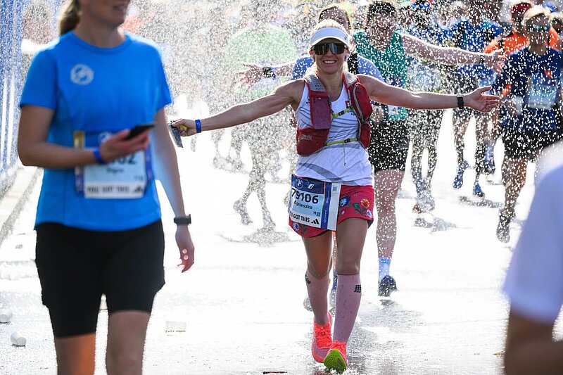 Läuferin beim BERLIN-MARATHON mit Kopfbedeckung und Sonnenbrille lächelt während sie durch Wasserregen läuft.