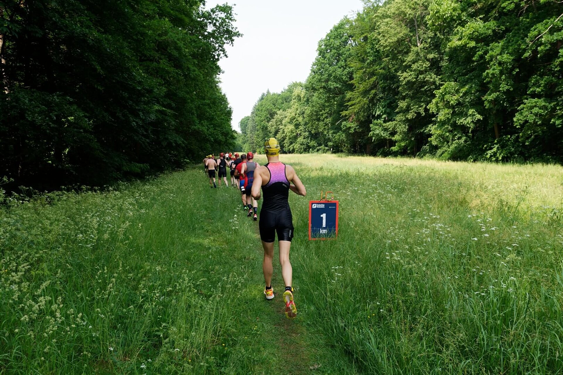 Läufer:innen in Neoprenanzügen und Badekappen passieren das 1-km-Schild auf einer von Bäumen umgebenen Wiese.