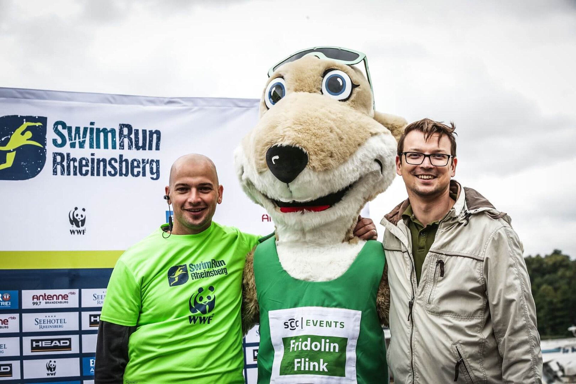 Michael Gerlach, Maskottchen Frido und Frank-Rudi Schwochow legen gegenseitig die Arme um die Schultern und lächeln in die Kamera.