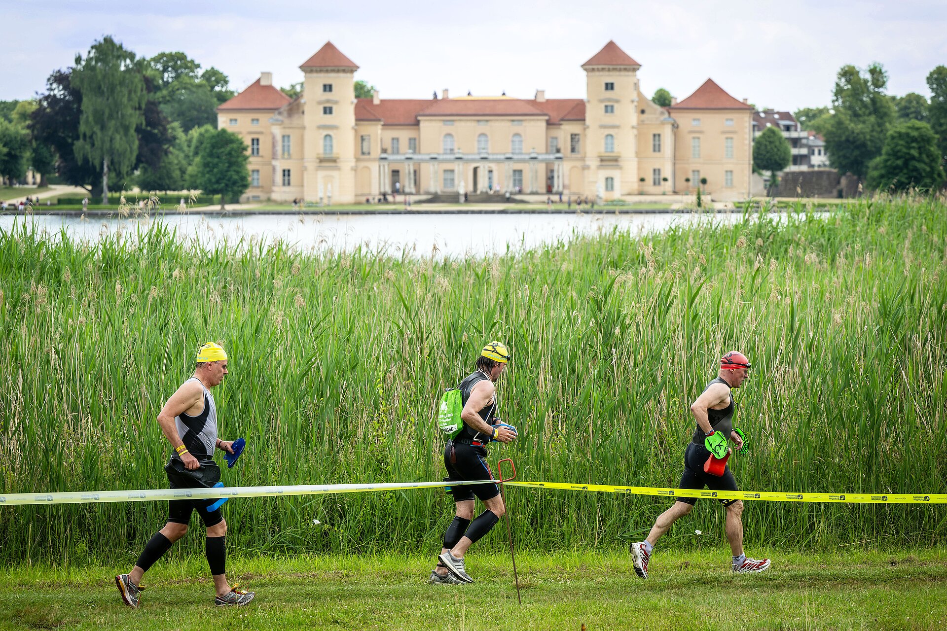 SwimRun: Three male participants run along the lake, Rheinsberg Castle in the background © SCC EVENTS / Tilo Wiedensohler