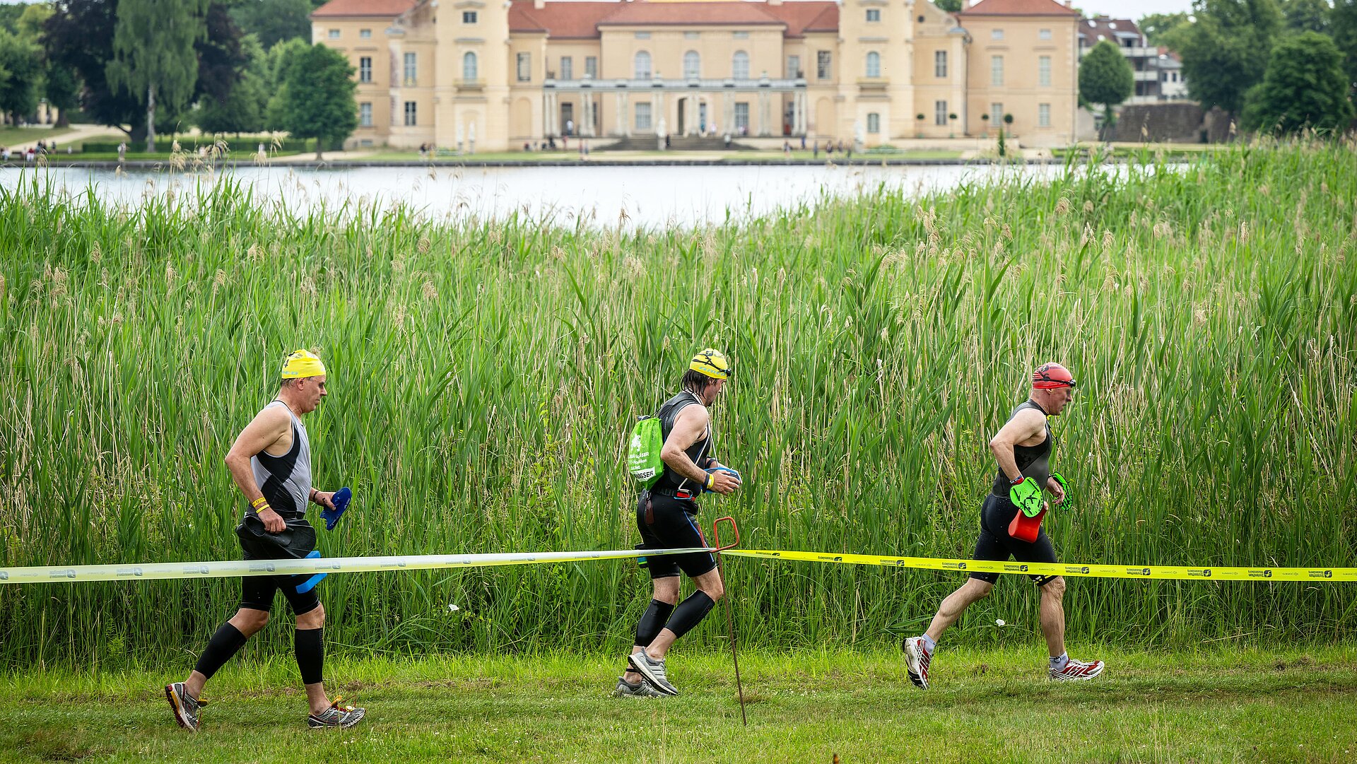 SwimRun: Three male participants run along the lake, Rheinsberg Castle in the background © SCC EVENTS / Tilo Wiedensohler