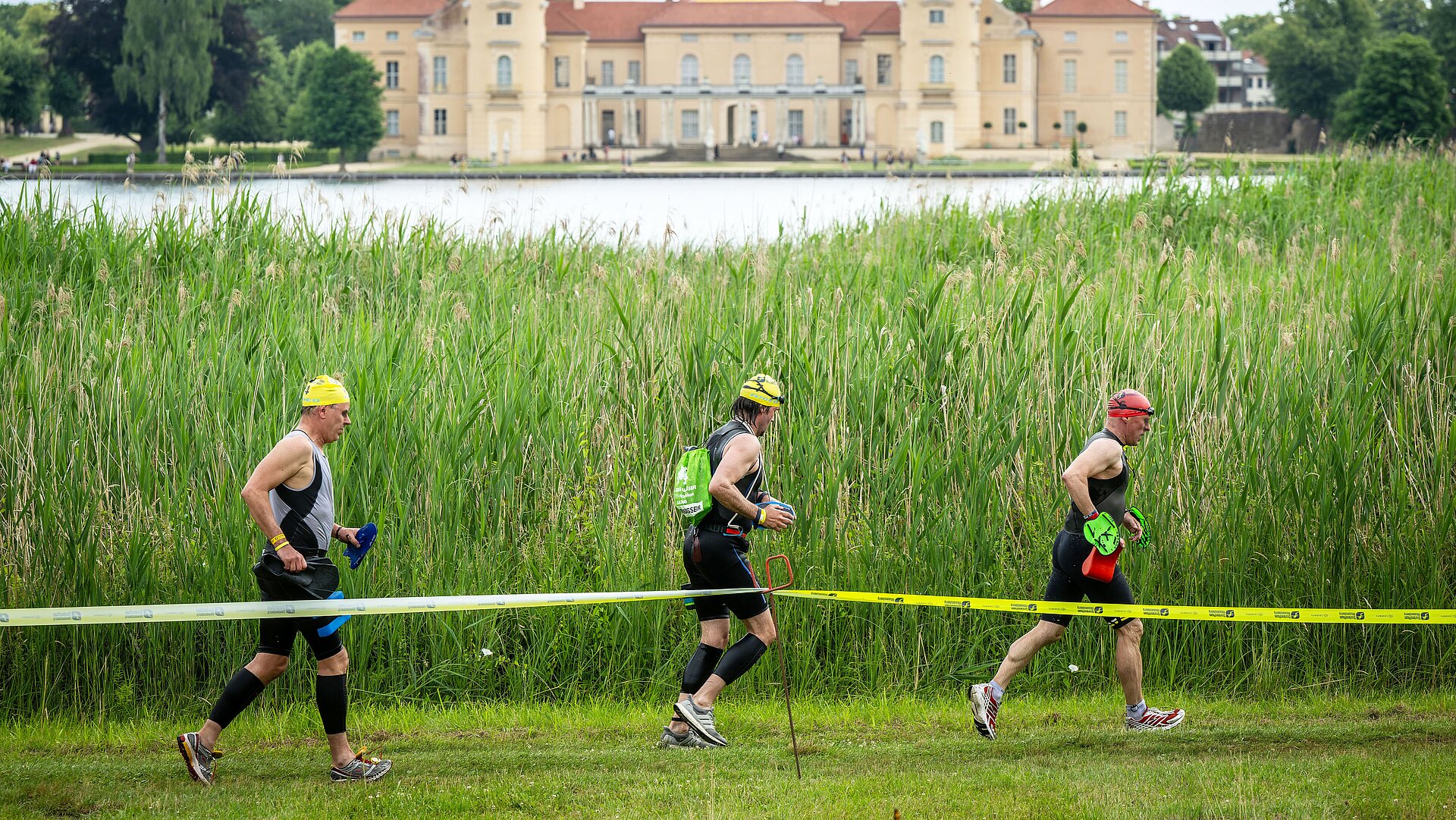 SwimRun: Drei Teilnehmer laufen am See entlang, Schloss Rheinsberg im Hintergrund © SCC EVENTS / Tilo Wiedensohler